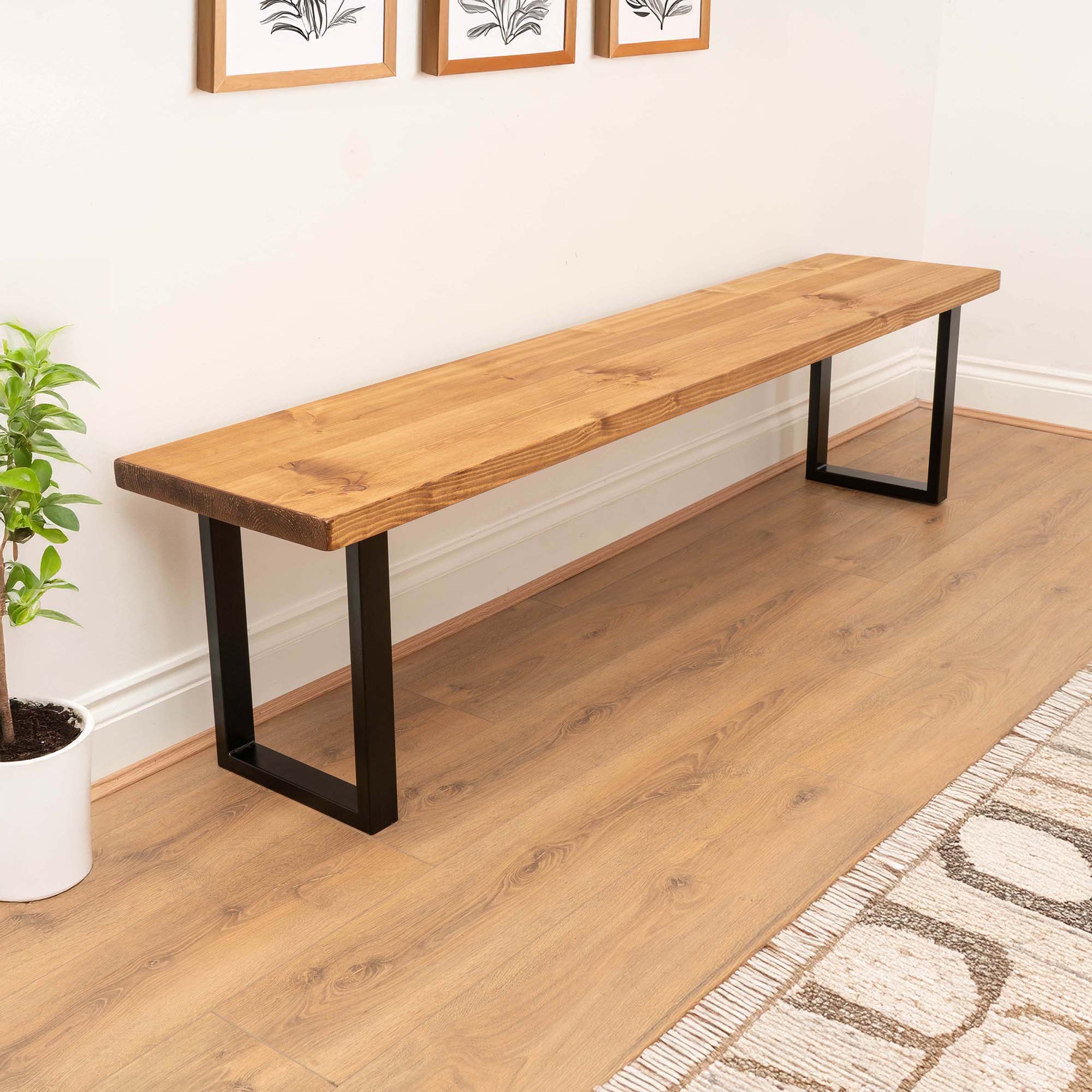 Wooden bench with black metal legs in a room with a plant and framed pictures on the wall.