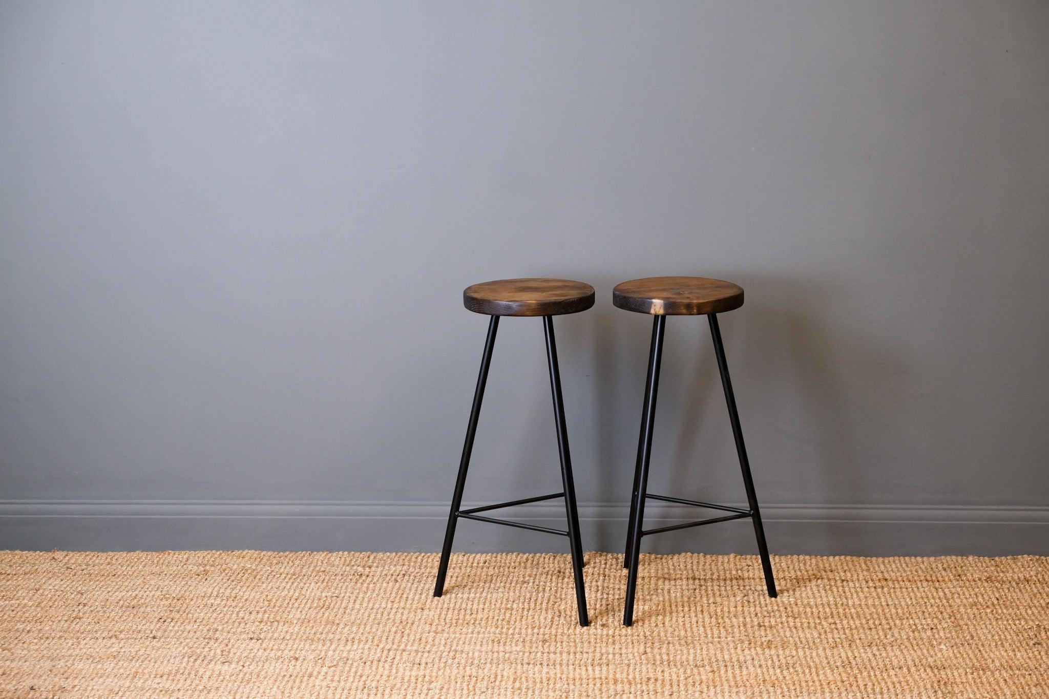 Two wooden stools with black metal legs against a gray wall.